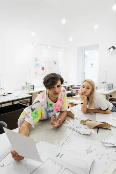 Stylish designer showing paper to colleague near sewing patterns — Stock Photo