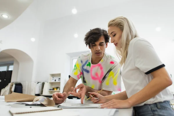 Fashion designer pointing with finger at paper near blonde colleague — Stock Photo