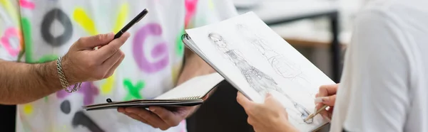 Partial view of fashion designer holding pen and notebook near drawing colleague, banner — Stock Photo