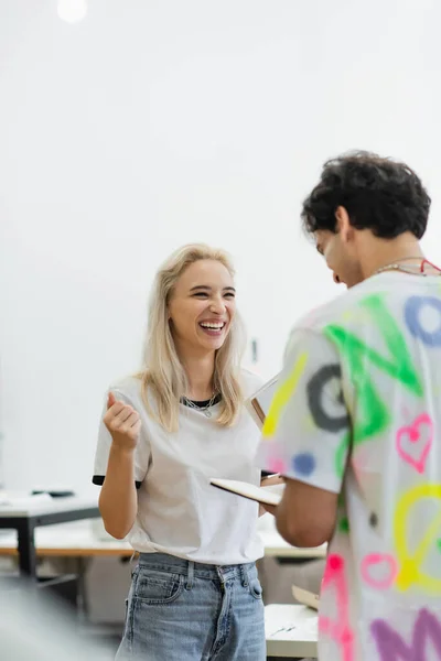 Excited fashion designer laughing near colleague with notebook in tailor shop — Stock Photo
