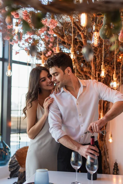 Mulher alegre abraçando namorado abrindo garrafa de vinho tinto no restaurante — Fotografia de Stock