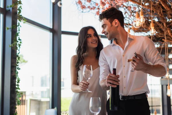 Uomo tenendo cavatappi con tappo vicino bottiglia e guardando allegra fidanzata in abito antiscivolo — Foto stock