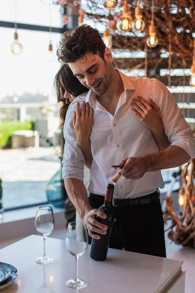 Mulher morena abraçando namorado feliz abrindo garrafa de vinho tinto no restaurante — Fotografia de Stock