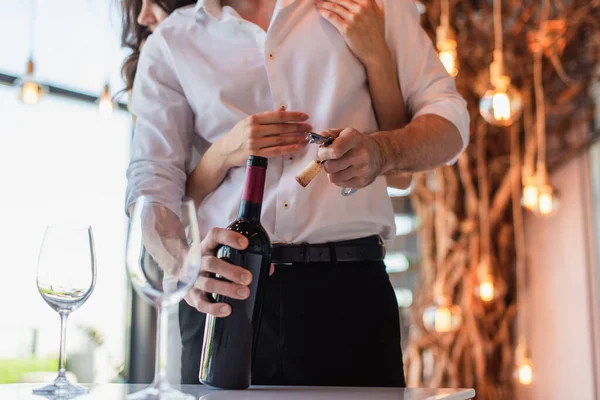 Vue recadrée de la femme brune étreignant petit ami ouverture bouteille de vin rouge dans le restaurant — Photo de stock