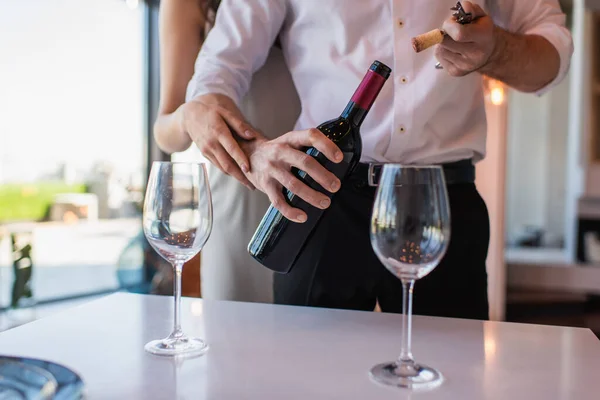 Vista recortada de la mujer abrazando novio apertura botella de vino tinto en el restaurante - foto de stock