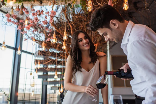 Homme versant du vin rouge dans un verre près de femme heureuse en robe glissante — Photo de stock