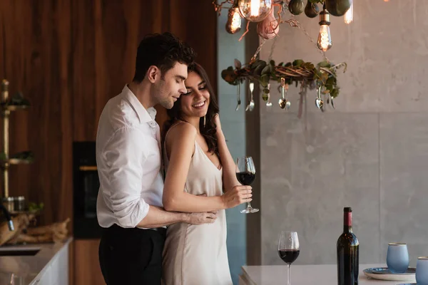 Homem abraçando feliz namorada em vestido de deslizamento com copo de vinho — Fotografia de Stock