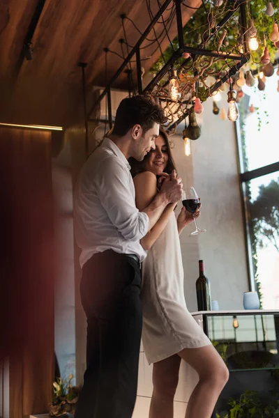 Cheerful man hugging happy girlfriend in slip dress with glass of wine — Stock Photo