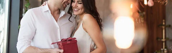 Homem feliz segurando caixa de presente vermelho perto de namorada sorridente em vestido de deslizamento, banner — Fotografia de Stock