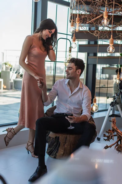 Homem feliz sentado com copo de vinho tinto e segurando a mão com a mulher em vestido de deslizamento — Fotografia de Stock
