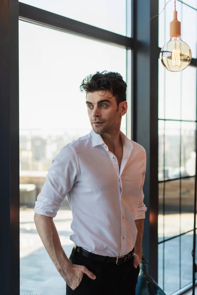 Elegante hombre de camisa blanca de pie con las manos en los bolsillos cerca de la ventana - foto de stock