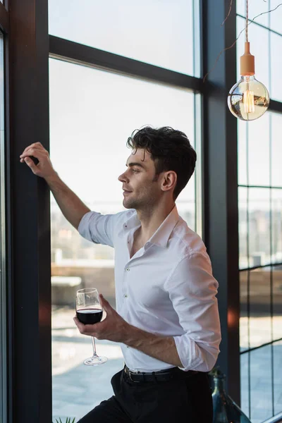 Elegante uomo in camicia bianca che regge un bicchiere di vino rosso in piedi vicino alla finestra — Foto stock