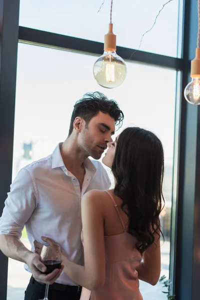 Homem segurando copo de vinho tinto e beijando mulher morena em vestido de deslizamento — Fotografia de Stock