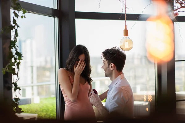 Hombre feliz haciendo propuesta a novia en vestido de deslizamiento cubriendo la cara en el restaurante - foto de stock