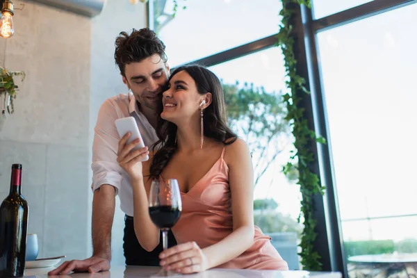 Casal feliz em fones de ouvido sem fio olhando uns para os outros perto de smartphone e garrafa de vinho — Fotografia de Stock