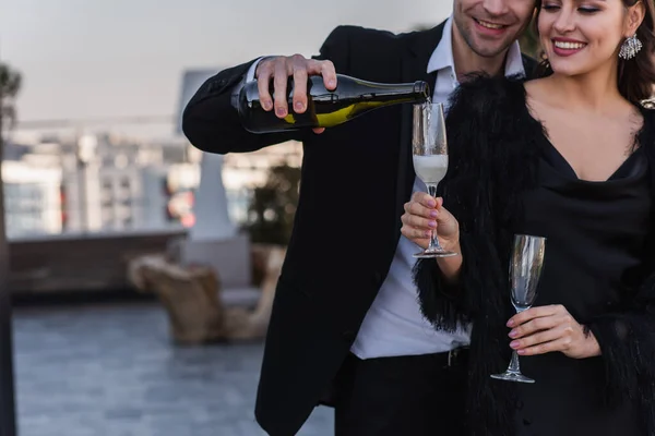 Happy man pouring champagne in glass of girlfriend in faux fur jacket — Stock Photo