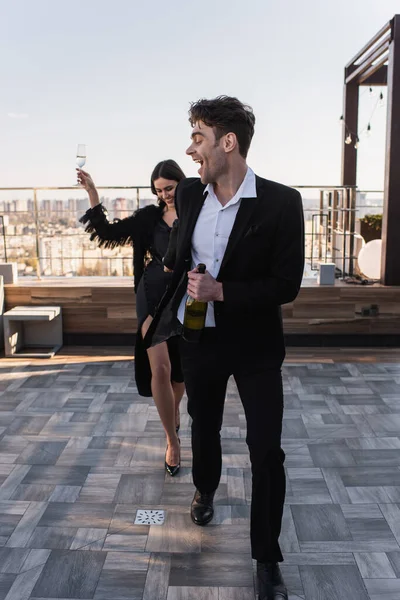 Повна довжина збудженого чоловіка тримає пляшку шампанського біля дівчини на терасі — стокове фото