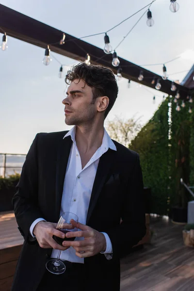 Elegant man in suit holding glass of red wine and looking way on terrace — Stock Photo