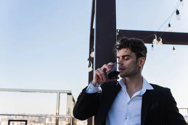 Elegante uomo in giacca e cravatta che tiene il bicchiere e beve vino rosso sul tetto — Foto stock