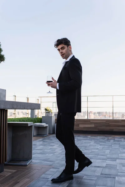 Longitud completa de hombre elegante en traje sosteniendo copa de vino tinto en la terraza - foto de stock