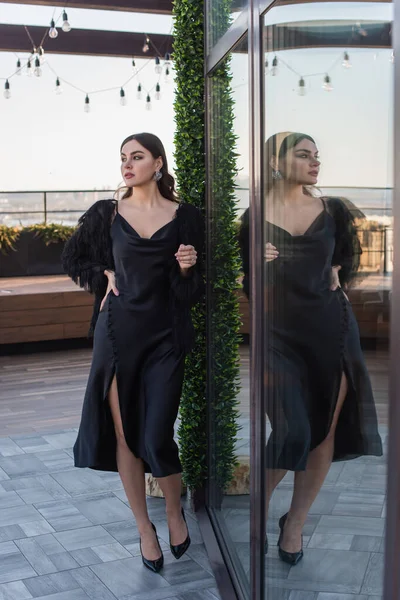 Volle Länge der jungen Frau in schwarzem Slip-Kleid und Kunstpelzjacke, die mit der Hand auf der Hüfte auf der Terrasse steht — Stockfoto