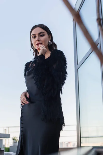 Vista de ángulo bajo de la mujer joven en vestido de deslizamiento negro y chaqueta de piel sintética en la terraza — Stock Photo