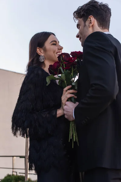 Hombre sosteniendo rosas rojas cerca de mujer feliz en chaqueta de piel sintética - foto de stock