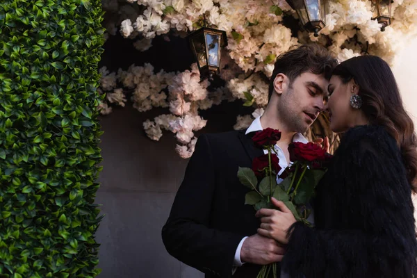 Man holding red roses near woman in faux fur jacket near blooming flowers on wall — Stock Photo