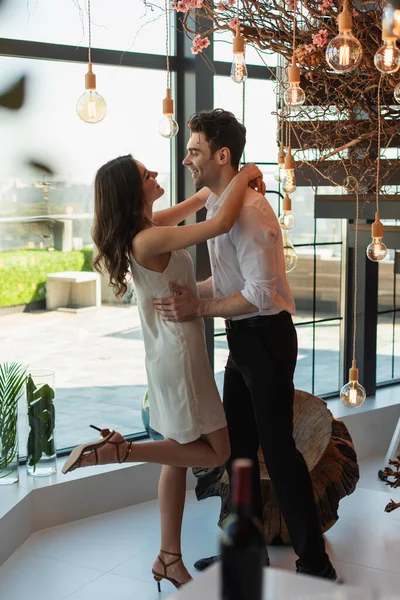 Side view of cheerful man in shirt hugging happy woman in slip dress in restaurant — Stock Photo