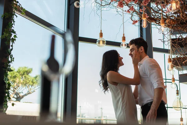 Vista lateral de la mujer feliz en vestido de deslizamiento tocando la cara del novio en el restaurante - foto de stock