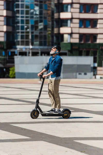 Full length of happy man in sunglasses and helmet riding e-scooter in urban city - foto de stock
