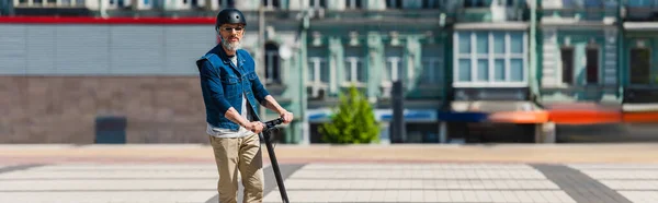 Зрілий чоловік в сонцезахисних окулярах і шоломі їде на електронному скутері в міському місті, банер — стокове фото