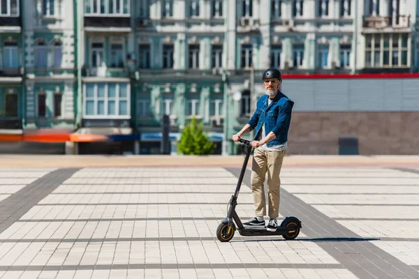 Full length of middle aged man in sunglasses and helmet riding e-scooter in urban city — Stock Photo