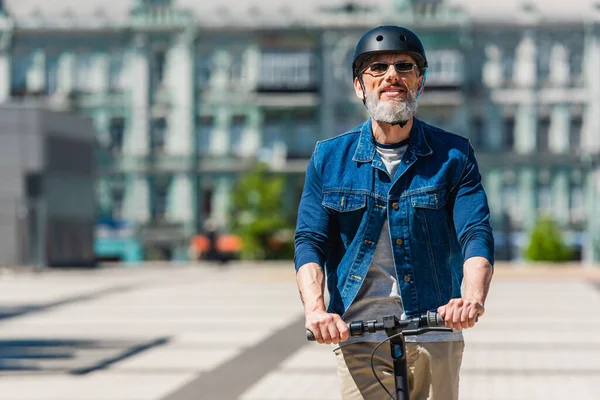 Pleased mature man in sunglasses and helmet riding e-scooter in urban city — Stock Photo