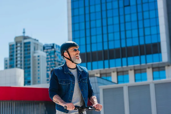 Positive and mature man in helmet and sunglasses riding electric scooter on street in city - foto de stock