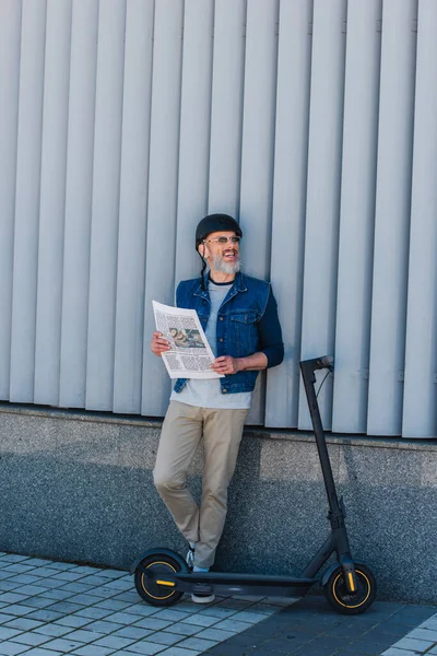 Full length of cheerful and mature hipster in helmet and sunglasses holding newspaper near e-scooter — Fotografia de Stock