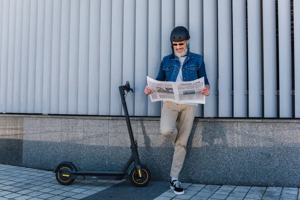 Full length of happy and mature hipster in helmet and sunglasses reading travel life newspaper near e-scooter — Stock Photo