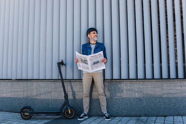 Full length of mature hipster in helmet and sunglasses holding travel life newspaper near e-scooter — Foto stock