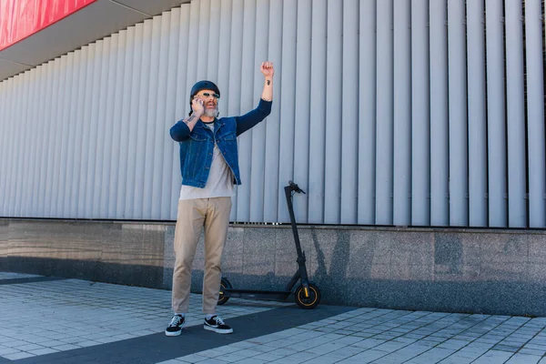 Full length of happy and mature hipster in helmet and sunglasses talking on smartphone and gesturing near e-scooter — Fotografia de Stock