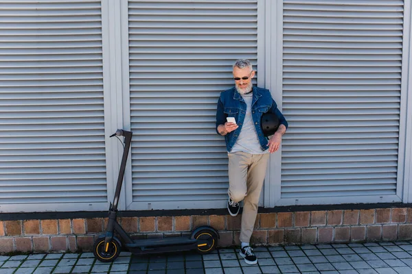 Full length of happy and mature man in sunglasses using smartphone near e-scooter — Stock Photo