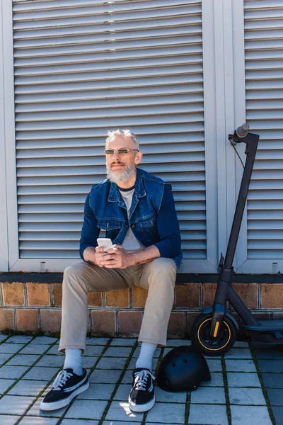 Full length of happy and mature man in sunglasses using smartphone and sitting near e-scooter — Stock Photo