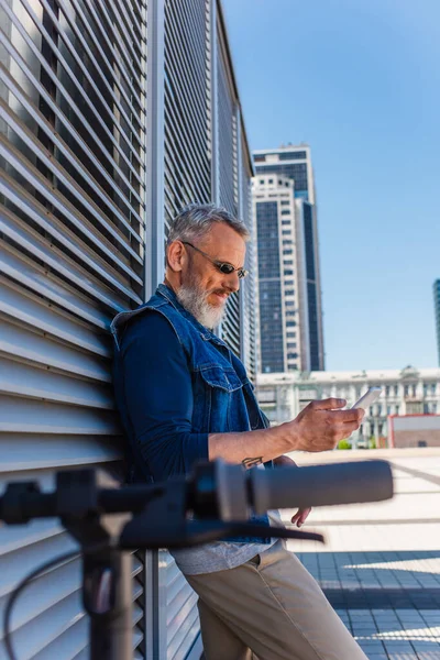 Mature man in sunglasses using smartphone near blurred e-scooter — Photo de stock