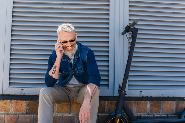 Fröhlicher und reifer Mann mit Sonnenbrille, der auf dem Smartphone spricht und in der Nähe von E-Scooter sitzt — Stockfoto