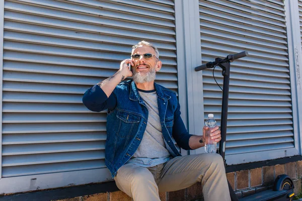 Fröhlicher und reifer Mann mit Sonnenbrille, der mit dem Smartphone spricht und eine Wasserflasche in der Nähe des E-Scooters hält — Stockfoto