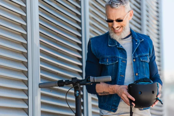 Бородатый мужчина средних лет в солнечных очках улыбается и держит шлем рядом с электрическим скутером — стоковое фото