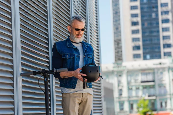 Middle aged man in sunglasses smiling and holding helmet near electric scooter — Foto stock