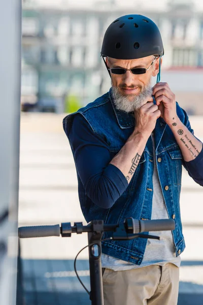 Tattooed middle aged man in sunglasses wearing helmet near electric scooter - foto de stock