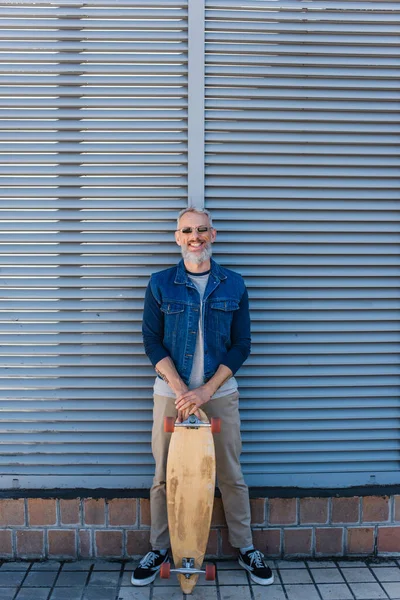 Full length of happy middle aged man in sunglasses holding longboard and looking at camera on urban street — Stock Photo