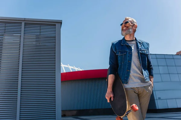 Низкоугольный вид позитивного мужчины средних лет в солнцезащитных очках, держащего лонгборд на городской улице — стоковое фото