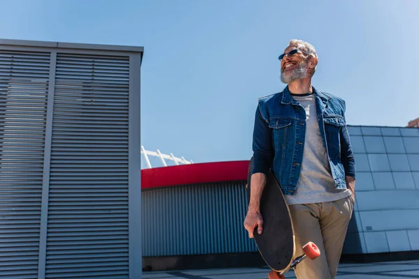 Low angle view of positive middle aged man in sunglasses holding longboard on urban street — Stock Photo